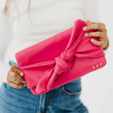 A person holding a pink leather clutch bag with a bow front and three gold stud details.